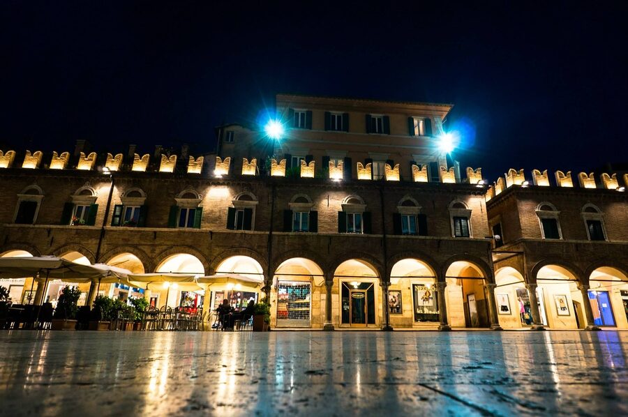 Ascoli Piceno's Piazza del Popolo at night with lit arcades and reflective wet travertine paving